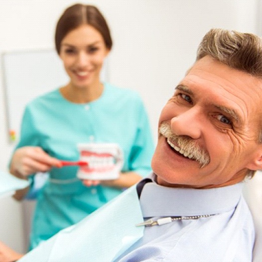 Senior man in dental chair smiling next to dental assistant brushing dentures
